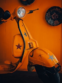 A retro-style yellow scooter adorned with star decals is positioned against an orange background. The scooter features a prominent headlight, black handles, and classic design elements. Woven baskets hang on the wall around it, adding to the vintage aesthetic.