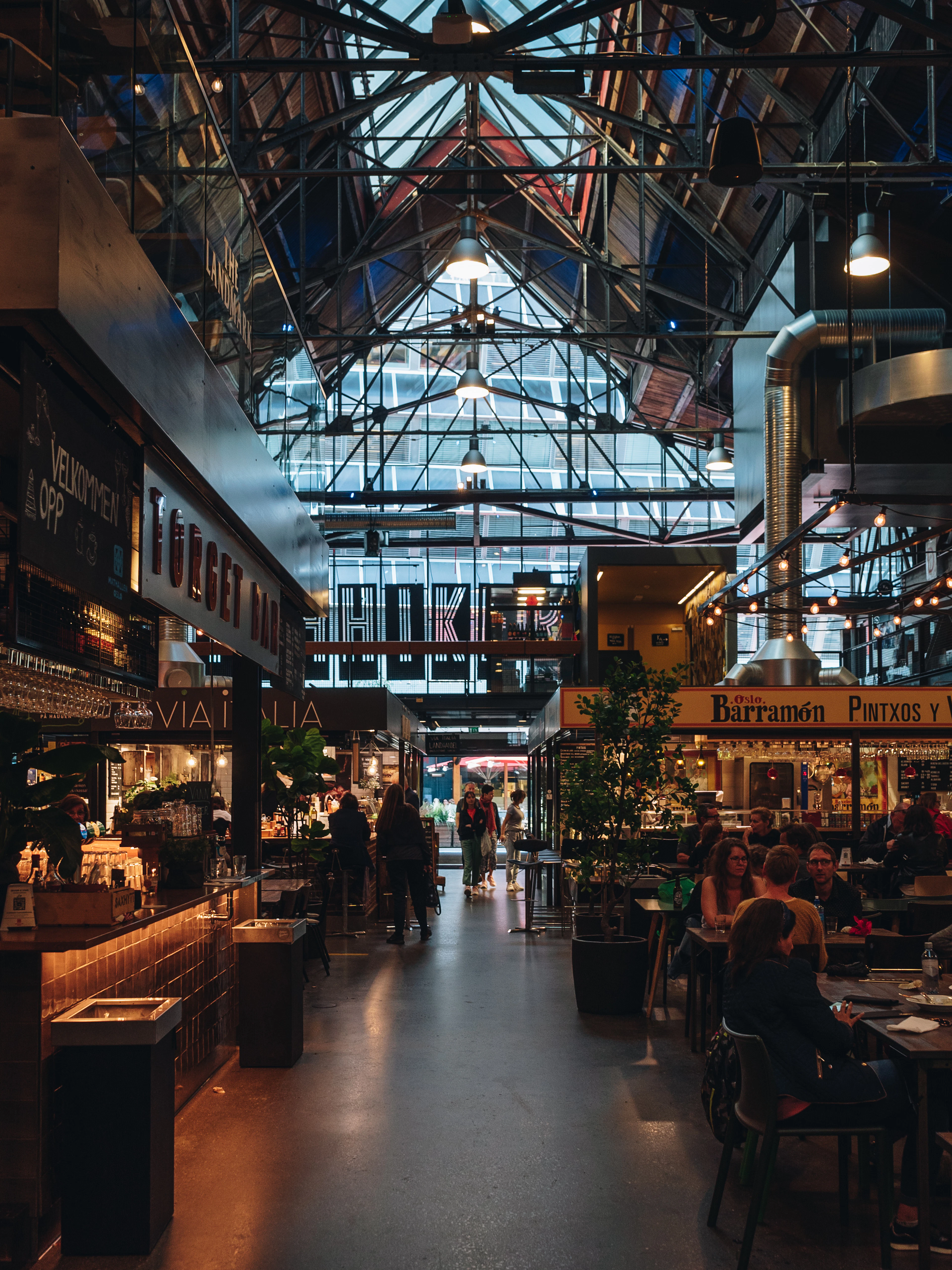 The glass-roofed hall of Mathallen food market in Oslo at dusk, lit stalls and counters running down both sides of the aisle beneath iron roof trusses while diners eat at long tables.