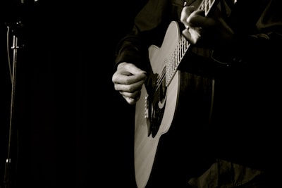 A person is playing an acoustic guitar with a focus on the hands strumming the strings. The background is dimly lit, creating a moody and intimate atmosphere.