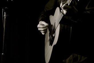 A person is playing an acoustic guitar with a focus on the hands strumming the strings. The background is dimly lit, creating a moody and intimate atmosphere.