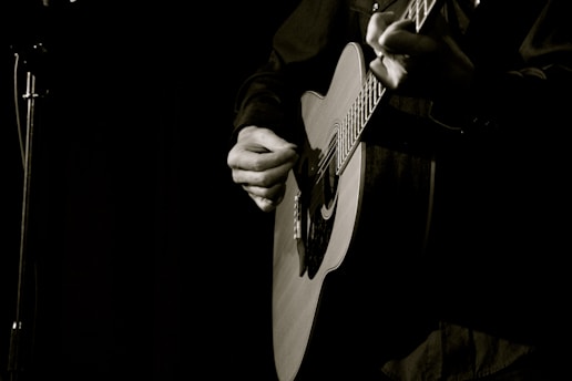 A person is playing an acoustic guitar with a focus on the hands strumming the strings. The background is dimly lit, creating a moody and intimate atmosphere.