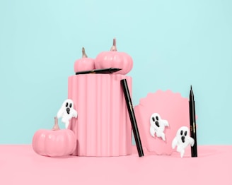 Pastel pink pumpkins are arranged creatively alongside a scalloped frame and black eyeliner pens. White ghost decorations add a whimsical and spooky touch against the soft blue background.