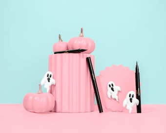 Pastel pink pumpkins are arranged creatively alongside a scalloped frame and black eyeliner pens. White ghost decorations add a whimsical and spooky touch against the soft blue background.