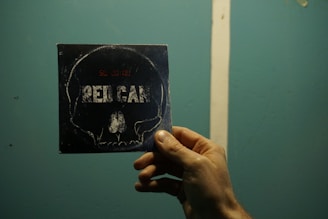 A hand holds a dark-colored card or CD case with an image of a skull and the text 'RED CAR' printed on it. The background is a solid turquoise color with a white vertical line.