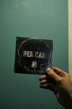 A hand holding a square card with a stylized skull outline and the word 'RED CAR' on it, set against a textured blue-green background.