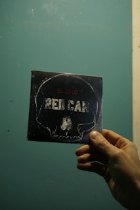 A hand holding a square card with a stylized skull outline and the word 'RED CAR' on it, set against a textured blue-green background.