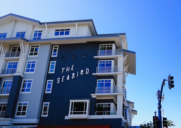 A modern coastal hotel building with a mixture of dark blue and white exterior. The building features multiple balconies with railings and large windows. The sign on the building reads 'The Seabird.' The sky is clear and blue, contributing to a serene atmosphere.