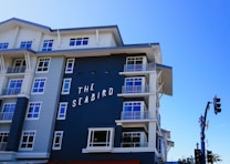 A modern coastal hotel building with a mixture of dark blue and white exterior. The building features multiple balconies with railings and large windows. The sign on the building reads 'The Seabird.' The sky is clear and blue, contributing to a serene atmosphere.