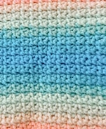 A close-up view of a crocheted fabric with a pattern of alternating colors. The texture shows a consistent and repetitive stitching pattern, transitioning smoothly from light blue to various shades of green and finally to peach.