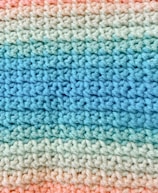 A close-up view of a crocheted fabric with a pattern of alternating colors. The texture shows a consistent and repetitive stitching pattern, transitioning smoothly from light blue to various shades of green and finally to peach.