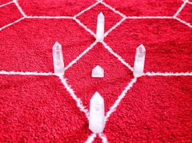 Clear quartz crystals arranged on a red rug with a white geometric pattern. The crystals are positioned in a symmetrical formation, creating a visually striking arrangement.