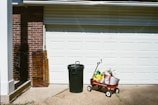 A garage door with a red wagon filled with bags of soil or fertilizer. Next to it stands a green trash can. Two pieces of stained glass rest against a brick wall to the left.