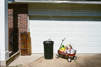 A garage door with a red wagon filled with bags of soil or fertilizer. Next to it stands a green trash can. Two pieces of stained glass rest against a brick wall to the left.