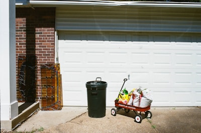 A garage door with a red wagon filled with bags of soil or fertilizer. Next to it stands a green trash can. Two pieces of stained glass rest against a brick wall to the left.