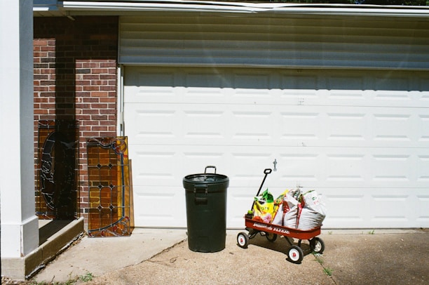 A garage door with a red wagon filled with bags of soil or fertilizer. Next to it stands a green trash can. Two pieces of stained glass rest against a brick wall to the left.