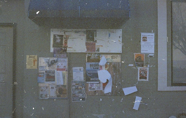 A weathered bulletin board filled with various posters and flyers against a gray wall. The posters advertise events, products, and announcements, some of which are peeling or partially obscured. A blue awning is visible above the board, and a window with a curtain is on the right side.