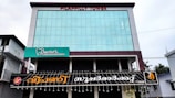 A modern building with large glass windows and a sign reading 'Pilappilly Tower' at the top. It houses a dentist clinic, with a green and white sign advertising it as a multi-specialty dental clinic. Below the clinic sign, there is a larger black sign with decorative text in another language. The surrounding environment includes electrical wires and a few trees visible to the right side.