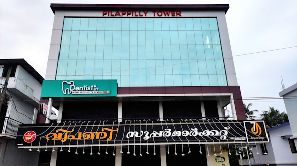 A modern building with large glass windows and a sign reading 'Pilappilly Tower' at the top. It houses a dentist clinic, with a green and white sign advertising it as a multi-specialty dental clinic. Below the clinic sign, there is a larger black sign with decorative text in another language. The surrounding environment includes electrical wires and a few trees visible to the right side.