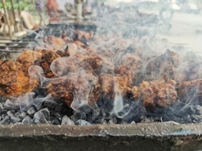 Chunks of meat are being grilled over hot charcoal, with smoke rising all around. The meat is browned and appears to be cooking on a barbecue grill. The background is slightly blurred, suggesting an outdoor setting with possible other grill setups.