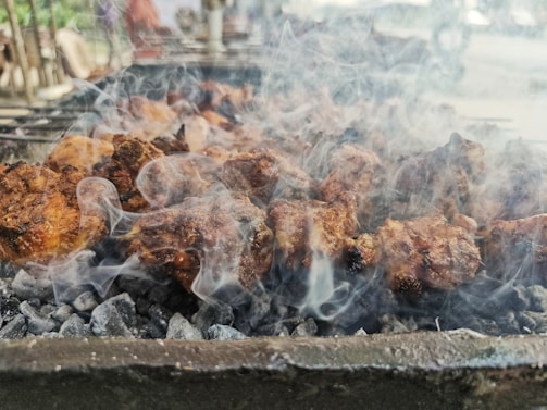 Chunks of meat are being grilled over hot charcoal, with smoke rising all around. The meat is browned and appears to be cooking on a barbecue grill. The background is slightly blurred, suggesting an outdoor setting with possible other grill setups.