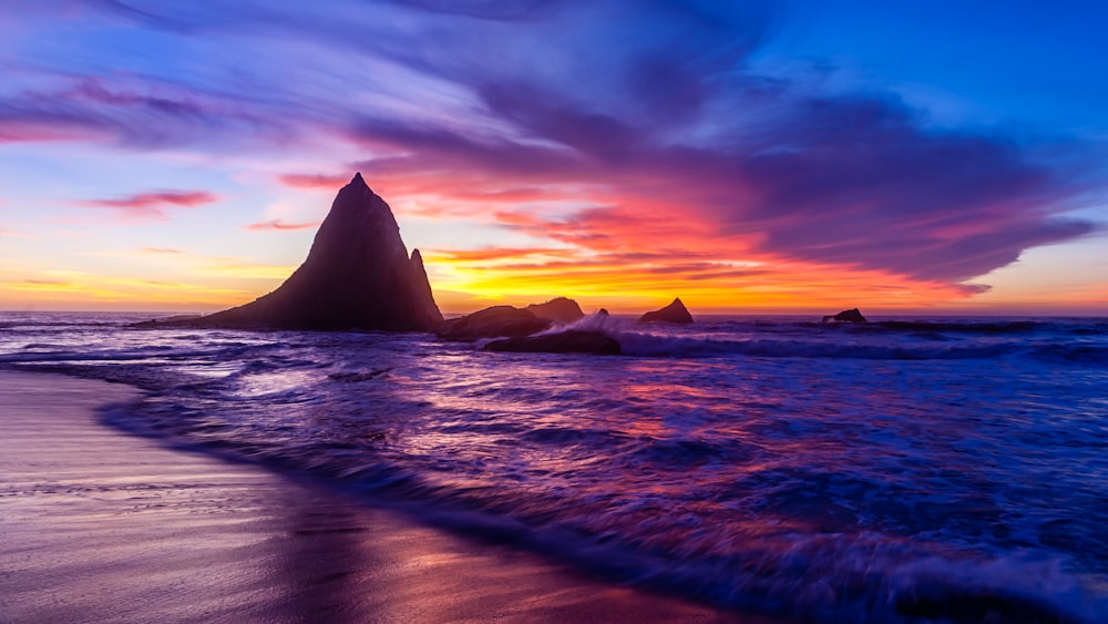 the sun sets over the ocean with a rock formation in the foreground