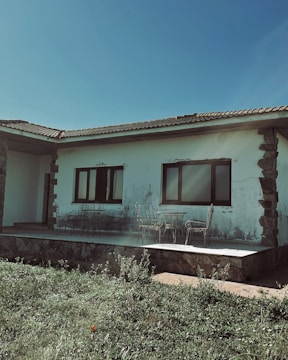 A single-story house with a tiled roof and stone accents on the corners. The building has two large windows and an outdoor patio area with wrought iron furniture, including chairs and a table. The lawn in the foreground is lush and green under a clear blue sky.