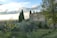 A rustic stone building with a tiled roof is surrounded by olive trees and tall cypress trees. The landscape includes grassy areas and scattered trees with a backdrop of a partly cloudy sky. The scene conveys a sense of tranquility and timeless countryside beauty.