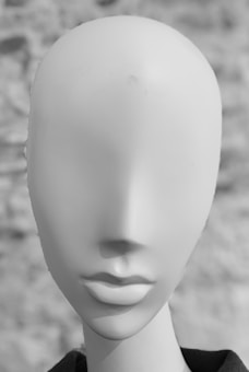 A smooth, featureless mannequin head against a blurred background. The head is sculpted to show a basic shape of a human face without any eyes, nose, or other facial features. The surface is smooth and matte, creating a minimalistic appearance.