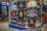 A display stands in a public area showcasing several images of athletes holding medals and flowers, presumably from the Olympics. The background includes people walking by and advertisements for smartphones.