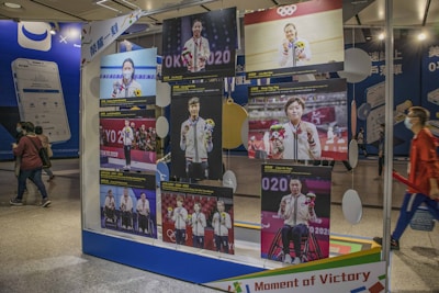 A display stands in a public area showcasing several images of athletes holding medals and flowers, presumably from the Olympics. The background includes people walking by and advertisements for smartphones.