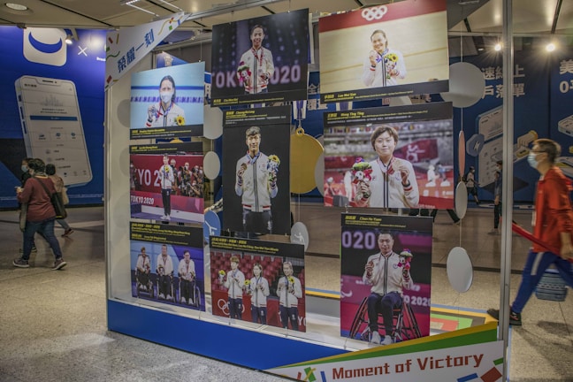 A display stands in a public area showcasing several images of athletes holding medals and flowers, presumably from the Olympics. The background includes people walking by and advertisements for smartphones.