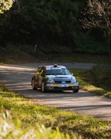 A rally car speeds along a winding road surrounded by lush greenery. The car is marked with sponsors and a racing number, indicating its participation in a motorsport event. Sunlight filters through the trees, casting patches of light and shadow on the road.