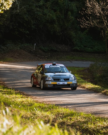 A rally car speeds along a winding road surrounded by lush greenery. The car is marked with sponsors and a racing number, indicating its participation in a motorsport event. Sunlight filters through the trees, casting patches of light and shadow on the road.
