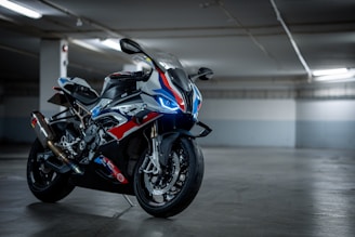 A sleek and modern sport motorcycle is parked in an indoor setting with subdued lighting. The bike features a dynamic design with a combination of blue, red, and white colors, and it has sharp, aerodynamic lines. It is equipped with performance-oriented components like disc brakes and a performance exhaust system. The setting appears to be a parking garage or similar structure, with a concrete floor and walls.
