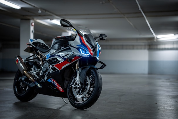 A sleek and modern sport motorcycle is parked in an indoor setting with subdued lighting. The bike features a dynamic design with a combination of blue, red, and white colors, and it has sharp, aerodynamic lines. It is equipped with performance-oriented components like disc brakes and a performance exhaust system. The setting appears to be a parking garage or similar structure, with a concrete floor and walls.