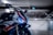 A sleek motorcycle with a prominent front view, featuring distinctive blue LED lights and aerodynamic design elements. The body is adorned with vibrant red, white, and blue graphics, and the motorcycle is situated in a dimly lit garage with industrial features in the background.