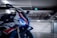A sleek motorcycle with a prominent front view, featuring distinctive blue LED lights and aerodynamic design elements. The body is adorned with vibrant red, white, and blue graphics, and the motorcycle is situated in a dimly lit garage with industrial features in the background.