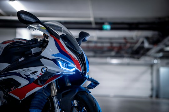 A sleek motorcycle with a prominent front view, featuring distinctive blue LED lights and aerodynamic design elements. The body is adorned with vibrant red, white, and blue graphics, and the motorcycle is situated in a dimly lit garage with industrial features in the background.