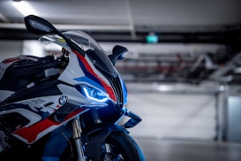 A sleek motorcycle with a prominent front view, featuring distinctive blue LED lights and aerodynamic design elements. The body is adorned with vibrant red, white, and blue graphics, and the motorcycle is situated in a dimly lit garage with industrial features in the background.