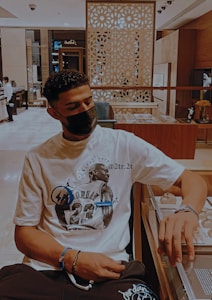 A person wearing a white graphic T-shirt with a prominent design related to basketball. They have a dark mask on and are seated in a modern, stylish interior with decorative patterns and display cases around them.