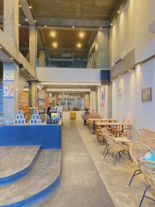 The interior of a modern, industrial-style caf&eacute; featuring high ceilings with exposed beams and sleek lighting. The space includes a counter with various products on display, wicker seating arrangements along the wall, and artwork adorning the walls. A mezzanine with glass barriers overlooks the main floor, adding depth to the environment. Patrons are seen sitting at tables, some working on laptops.
