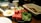 A breakfast spread featuring sunny-side-up eggs, sliced cucumbers, watermelon chunks, greens, walnuts, cheese, and flatbread. A white cup and glass jug of cream or milk are on the wooden table.