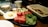 A breakfast spread featuring sunny-side-up eggs, sliced cucumbers, watermelon chunks, greens, walnuts, cheese, and flatbread. A white cup and glass jug of cream or milk are on the wooden table.