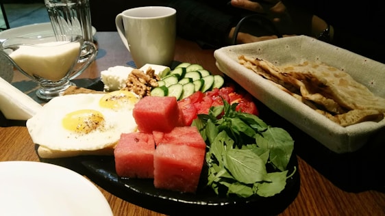 A breakfast spread featuring sunny-side-up eggs, sliced cucumbers, watermelon chunks, greens, walnuts, cheese, and flatbread. A white cup and glass jug of cream or milk are on the wooden table.