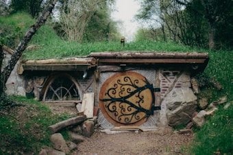 A rustic, small home is built into a gentle hillside, featuring a large, round wooden door adorned with decorative black wrought iron. The structure is partially covered by grass and earth, blending seamlessly with the surrounding greenery. The facade includes brick and stone elements, and the area is surrounded by trees and foliage.