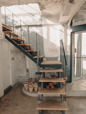 A rustic interior space featuring an industrial-style staircase with wooden steps and black metal railings. The walls are plastered with a mix of peeling paint and exposed concrete, adding to the raw aesthetic. On the floor, there are several terracotta pots, some with plants and others empty, arranged in a neat cluster. Natural light streams in, casting soft shadows across the textured surfaces.
