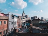 The urban landscape features a mix of residential buildings with a prominent church tower in the background. The structures vary in color and architectural style, with brick and concrete facades. A clear blue sky with scattered clouds provides a contrast to the man-made environment.
