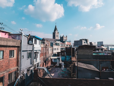 The urban landscape features a mix of residential buildings with a prominent church tower in the background. The structures vary in color and architectural style, with brick and concrete facades. A clear blue sky with scattered clouds provides a contrast to the man-made environment.