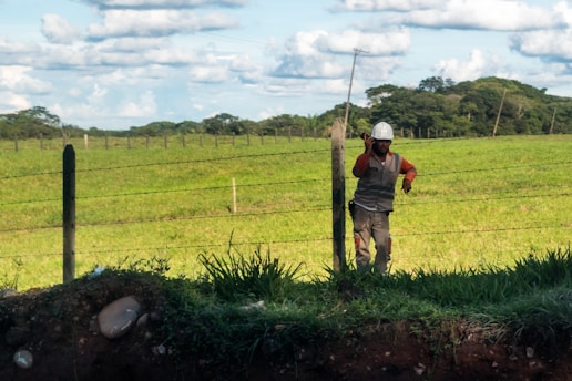 A person wearing a hard hat and protective clothing is standing near a fence in a grassy field. The person appears to be talking on a phone. The background shows a wide expanse of green grass with some trees in the distance and a partly cloudy blue sky.