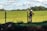 A person wearing a hard hat and protective clothing is standing near a fence in a grassy field. The person appears to be talking on a phone. The background shows a wide expanse of green grass with some trees in the distance and a partly cloudy blue sky.